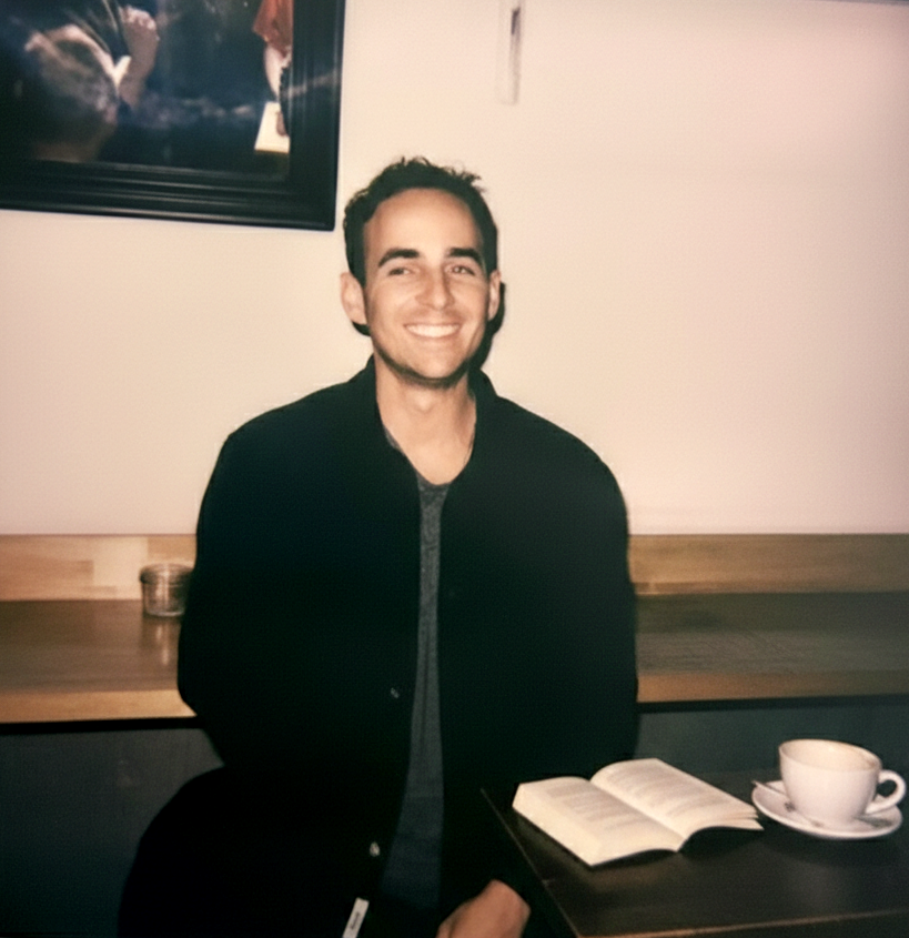 Jeffrey Masters, smiles at someone off camera. He is white with brown hair. A book and coffee cup are on the table in front of him.